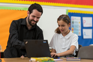 Male Teacher Helping Young School Girl How To Use Laptop